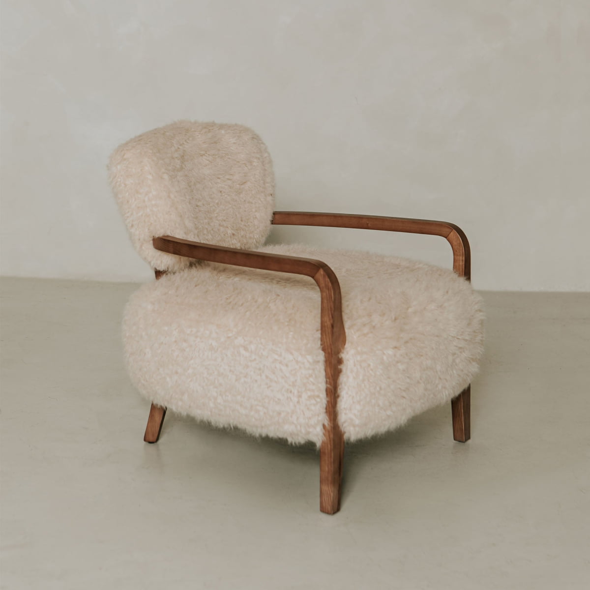 The Malmo Chair features minimalist elegance with a curved wooden frame and plush cream shearling upholstery, highlighted by a simple light-colored wall and floor.