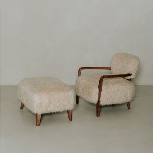 The Malmo Chair with Ottoman features wooden armrests and legs and is upholstered in cream shearling, creating minimalist sophistication against a light wall and floor.