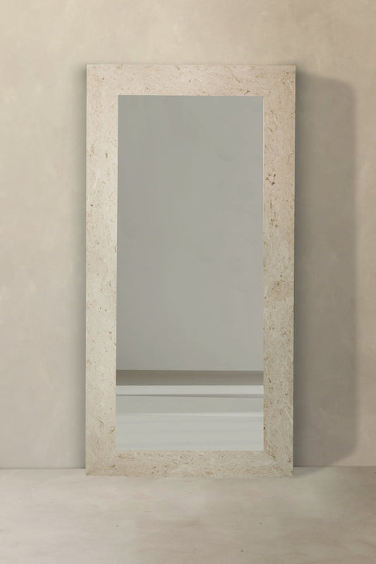 The Marbella Floor Mirror is a full-length rectangular mirror with a travertine stone frame, shown leaning against a textured light wall and reflecting a bare wall with a white baseboard on the smooth floor.