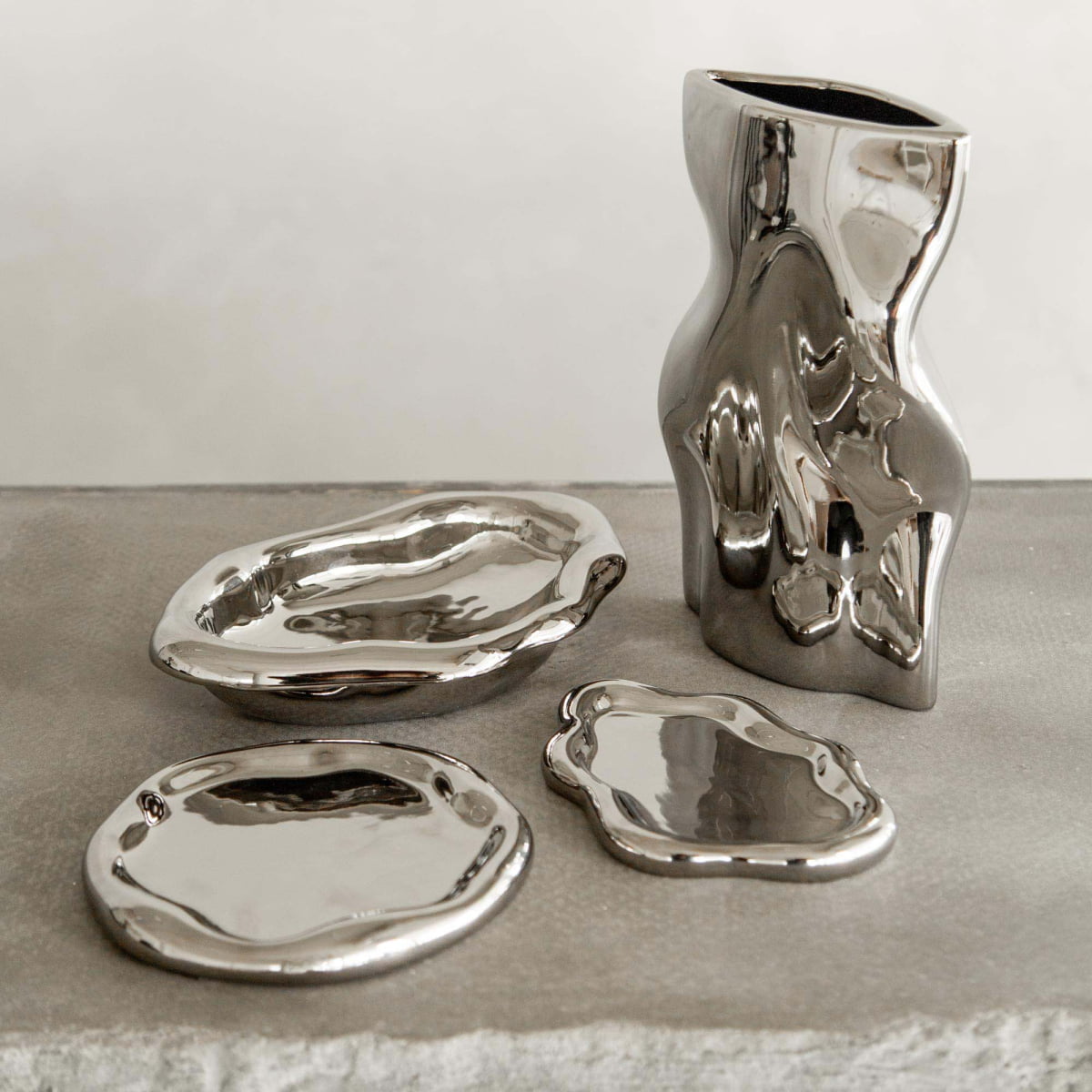 The Mira set features a polished chrome finish with one wavy-edged vase and three organically shaped plates resting on stone, showcasing modern sophistication with their glossy, reflective silver ceramic surfaces.