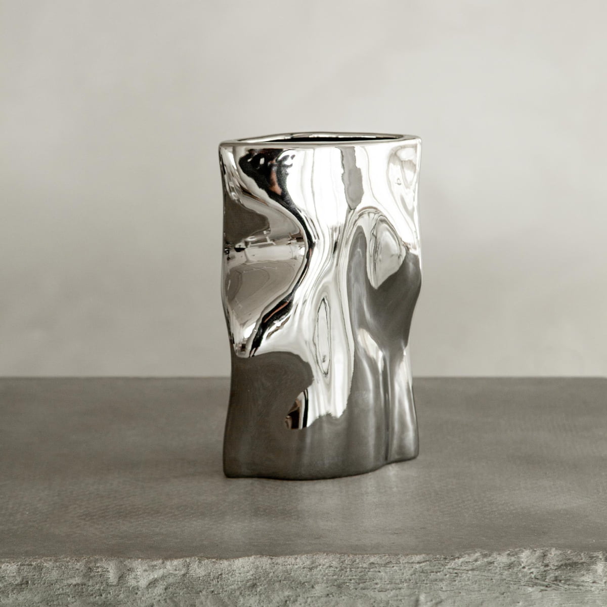 The Mira vase, with its polished chrome finish and abstract, organic shape, stands on a textured gray surface against a neutral backdrop, exuding modern sophistication.