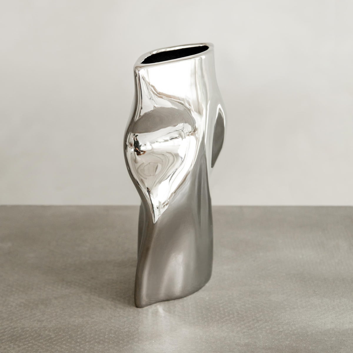 The Mira vase, featuring a polished chrome finish and organic abstract shape, sits on a textured gray surface against a light neutral background.