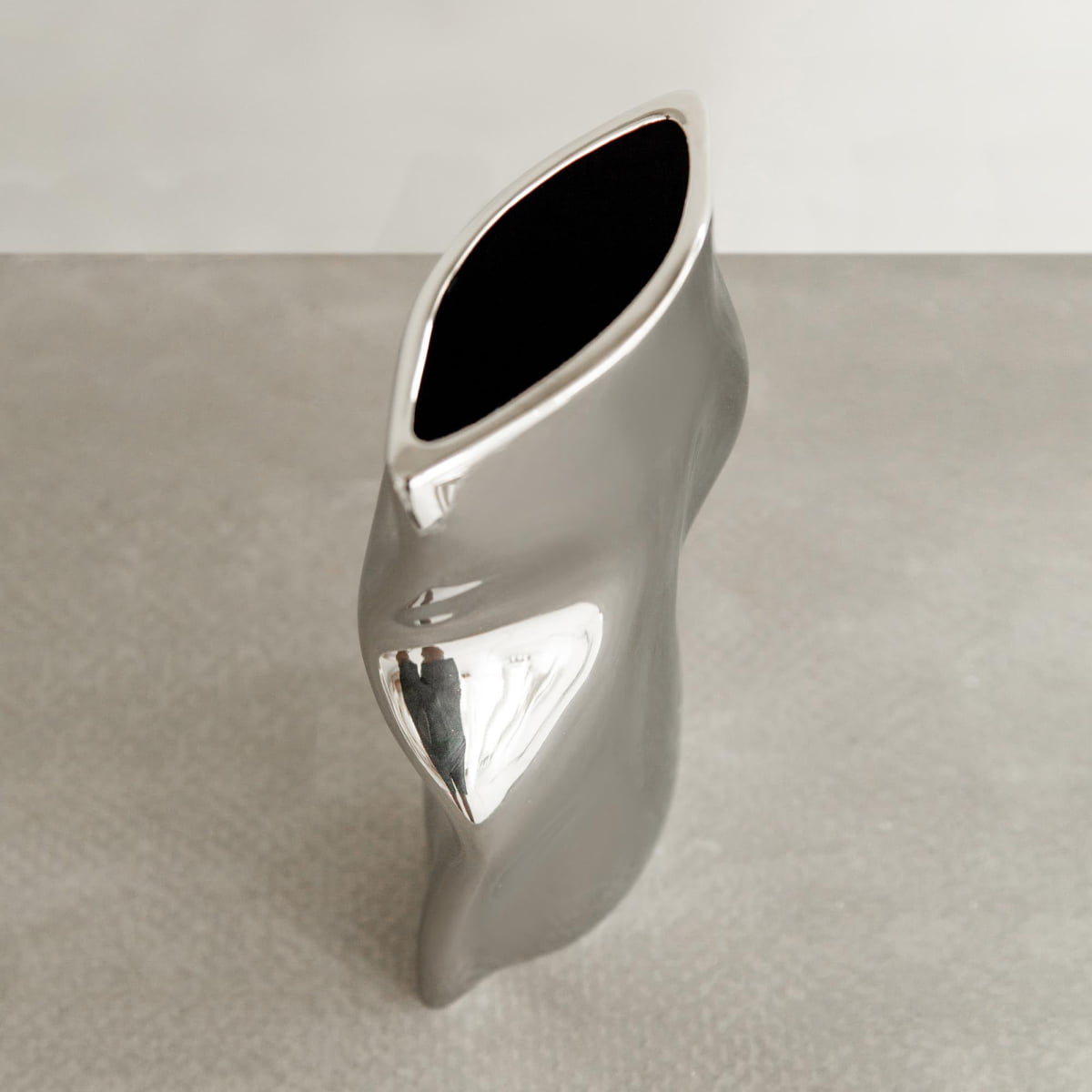 The Mira vase features a tall, modern silver design with a twisted, organic shape and wide oval opening, placed on a textured light gray surface for a touch of modern sophistication.