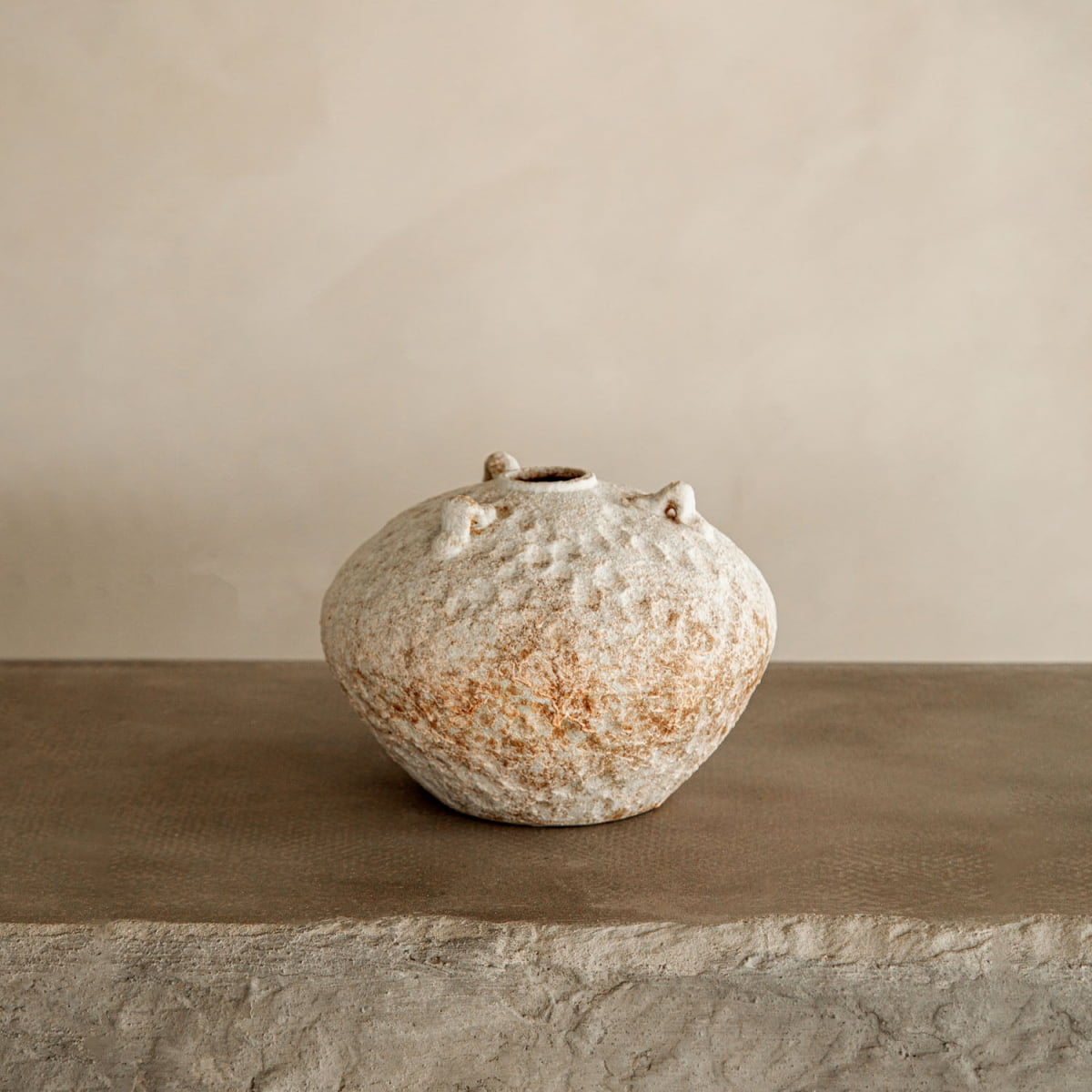 The Mizu vase, a rustic ceramic piece with a round shape and narrow opening, sits on stone against a beige background. Its earthy mottled brown and cream tones make this artisan pottery perfect for farmhouse decor.
