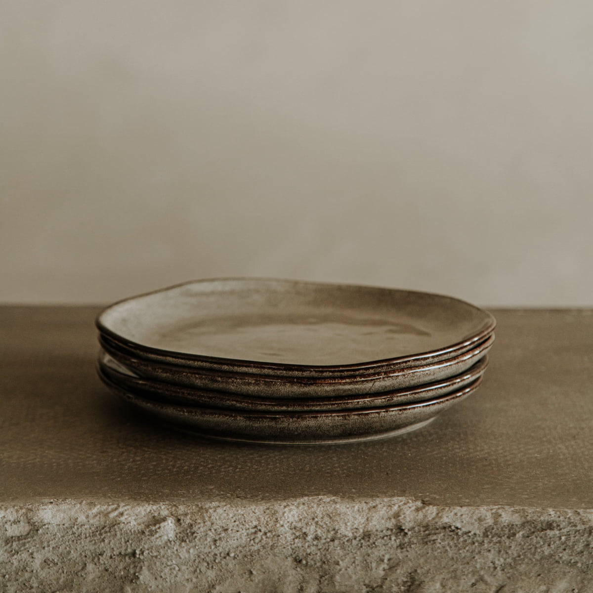 A stack of four rustic, uneven ceramic plates from the Nebula Stoneware Collection Dinnerplate Set sits on a textured stone surface, highlighting their earthy tones and stone-like finish against a neutral backdrop.