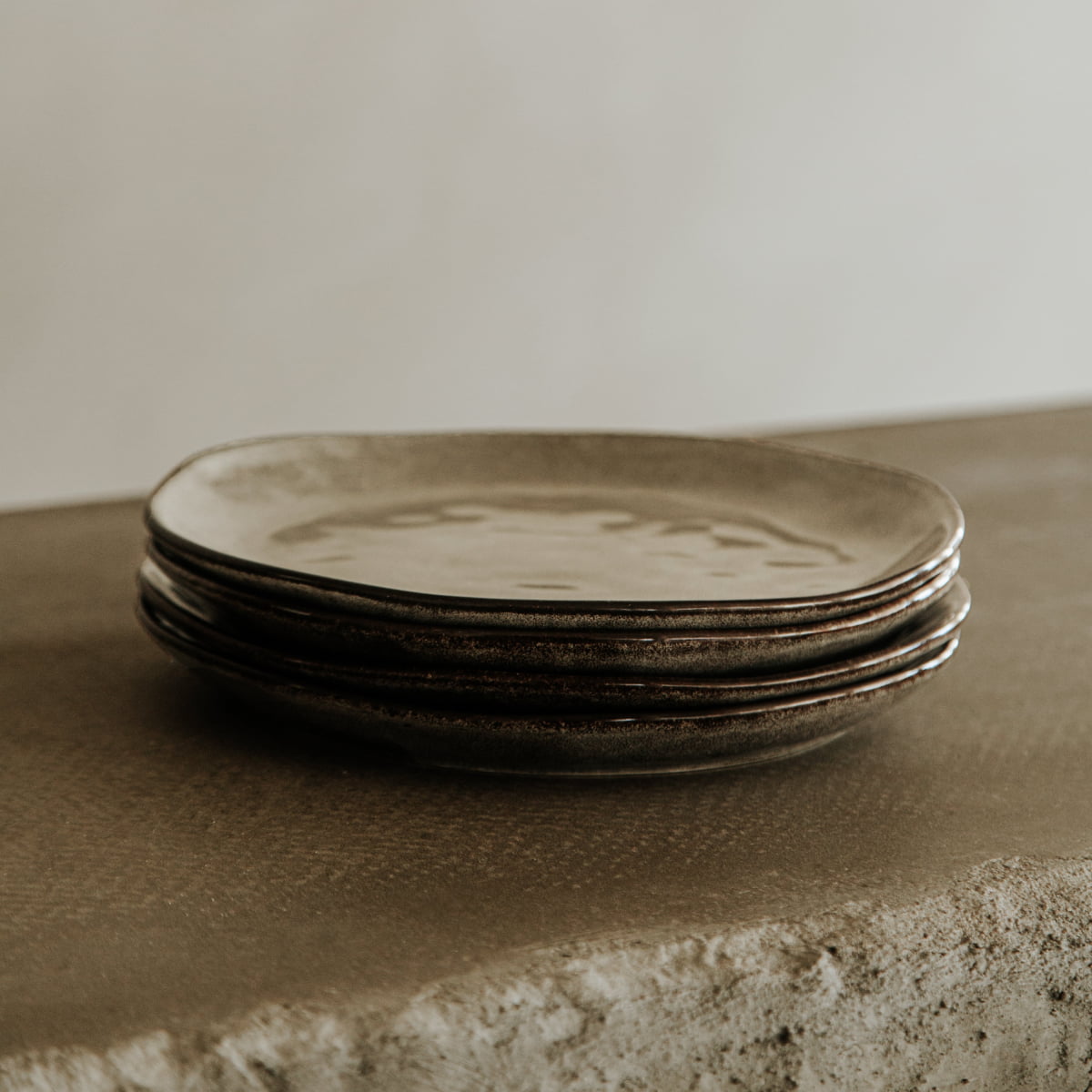 A stack of four irregularly shaped dinner plates from the Nebula Stoneware Collection Dinnerplate Set, featuring a rustic, stone-like finish, sits on a textured stone surface with a neutral, blurred background.