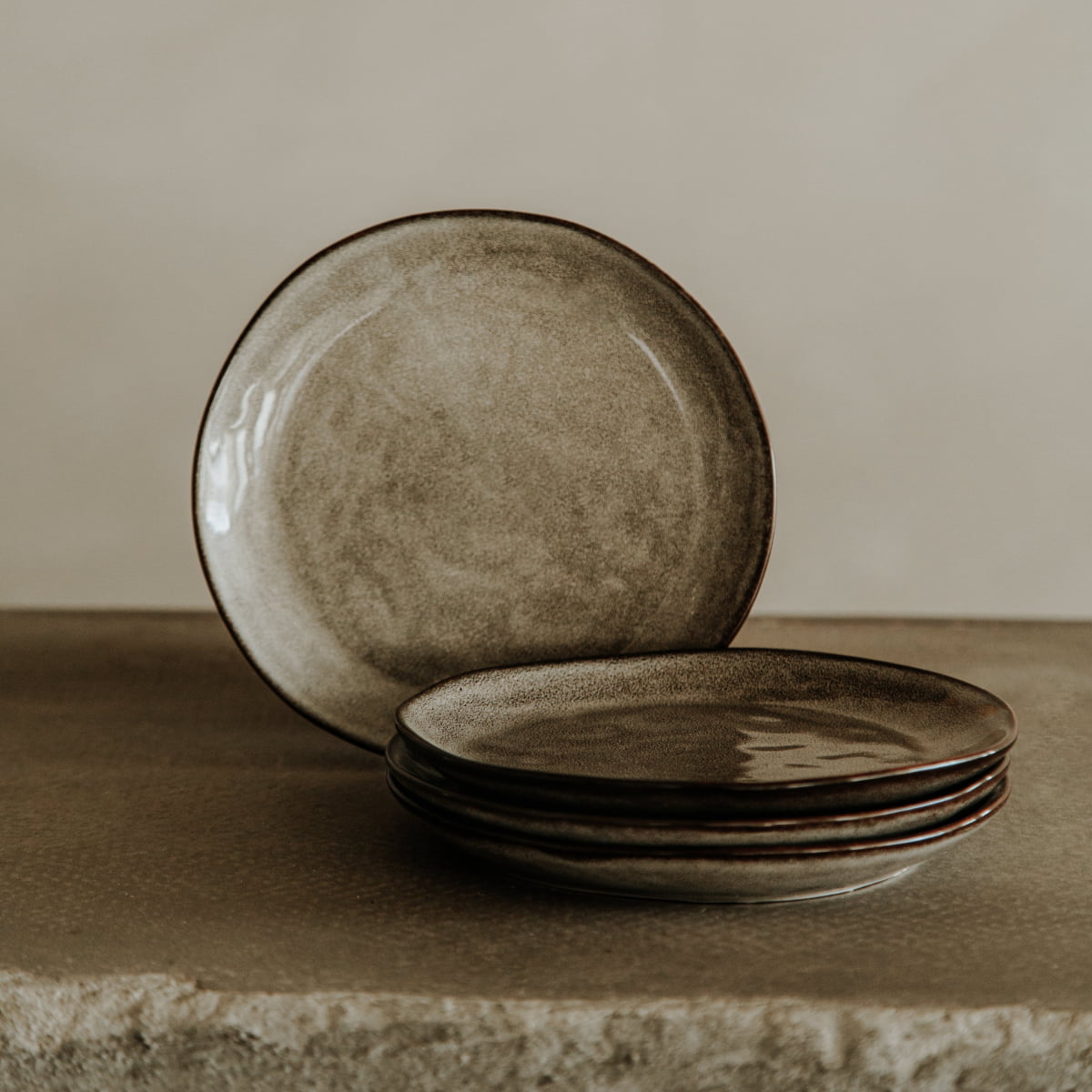 Four dinner plates from the Nebula Stoneware Collection Dinnerplate Set, featuring a stone-like finish and earthy tones, are stacked on a textured surface with one upright plate behind them against a softly lit neutral background.