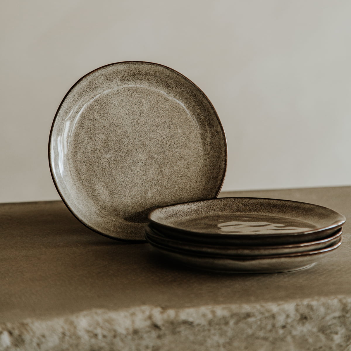 Four plates from the Nebula Stoneware Collection Dinnerplate Set, with a stone-like finish in earthy tones, are stacked on a textured surface, one upright behind them. The softly lit background is neutral.