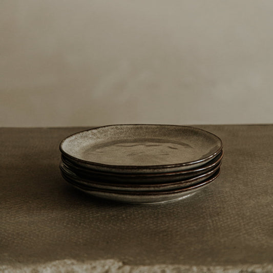 A set of four rustic dinner plates from the Nebula Stoneware Collection, with a matte stone-like finish, is stacked on a textured surface against a plain neutral background.