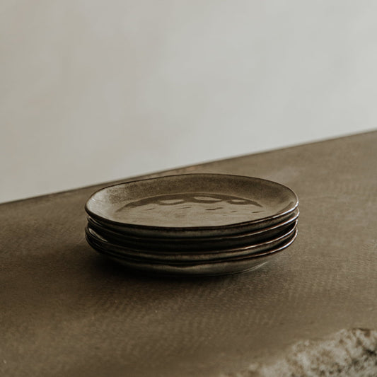 A stack of four rustic, dark Nebula Stoneware Collection Dinnerplate Set plates rests on a textured brown surface with a softly blurred neutral background.