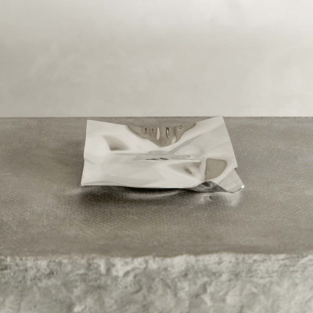 The Nexus Tray features a square design with polished chrome finish and crumpled edging, displayed on a textured light gray surface for a modern touch.