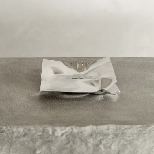 The Nexus Tray features a square design with polished chrome finish and crumpled edging, displayed on a textured light gray surface for a modern touch.