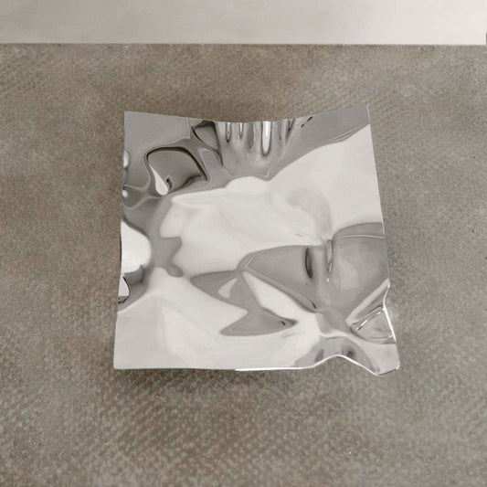 The Nexus Tray, featuring a square crumpled design and polished chrome finish, rests on a textured gray surface. Its abstract reflections and wavy contours create a bold contemporary look.