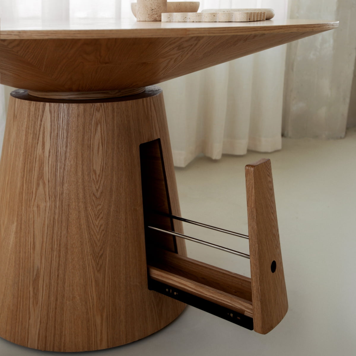 A close-up of the Nola + Elsa Dining Set shows its modern round wood table with a conical base featuring a hidden pull-out storage drawer, set on a smooth light floor with sheer white curtains in the background.
