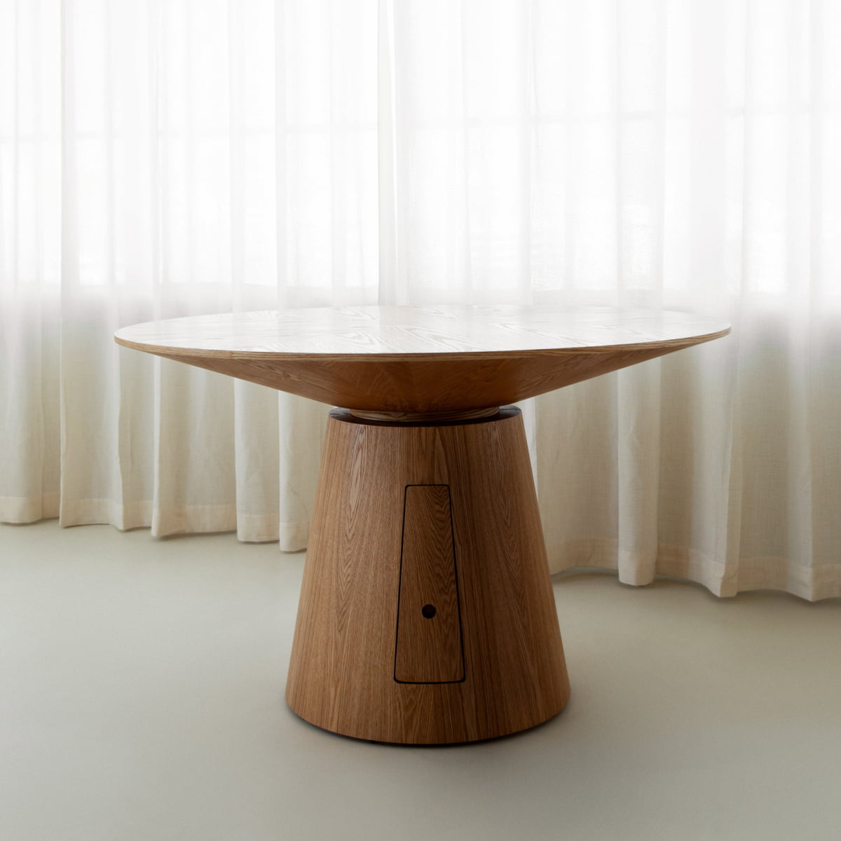 The Nola Dining Table features a thick, conical base and wide round top, displayed in front of sheer white curtains on a light floor—an elegant piece that highlights timeless craftsmanship.