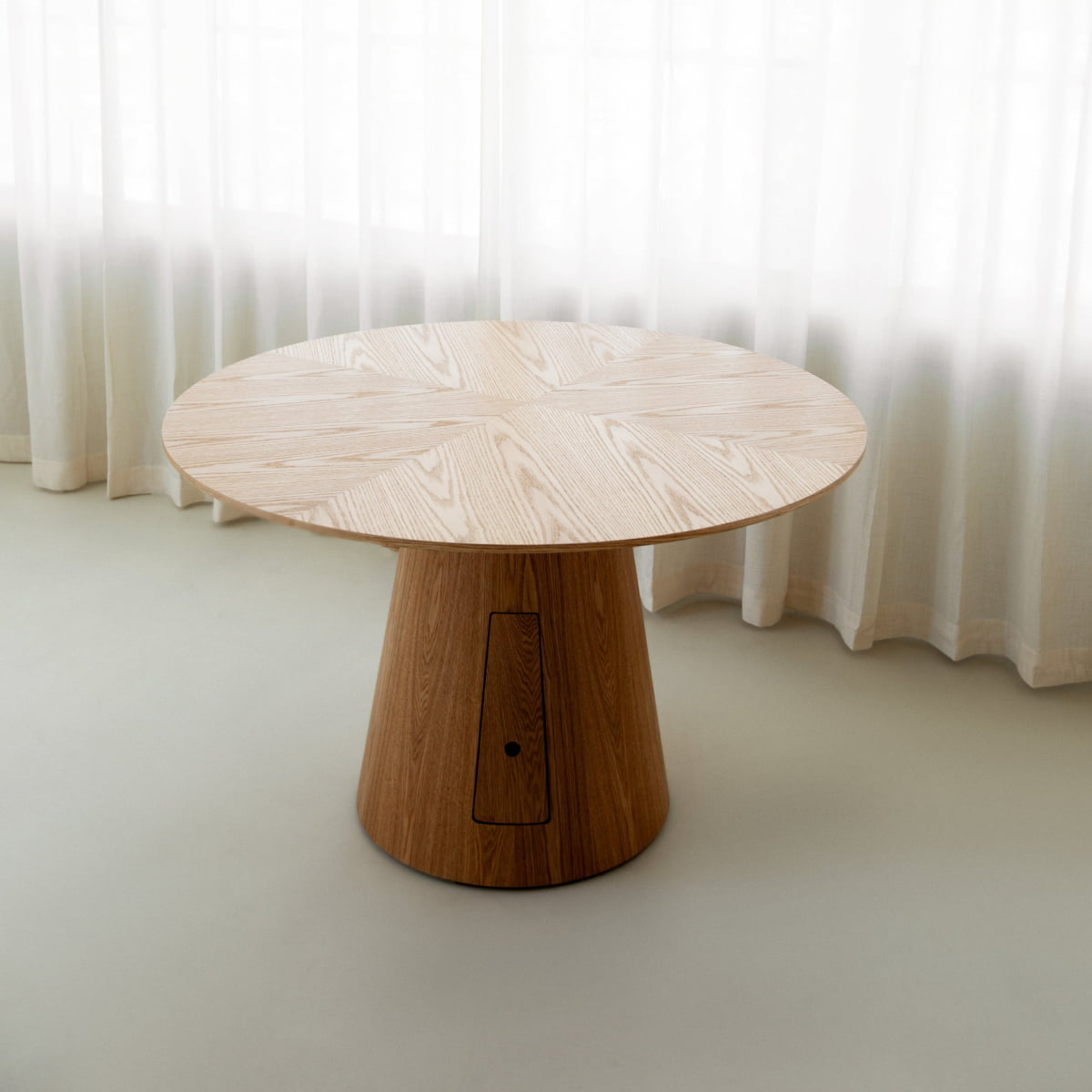 The Nola + Elsa Dining Set features a round wood table with a geometric patterned top and a wide, conical base, placed on a light floor before sheer white curtains.