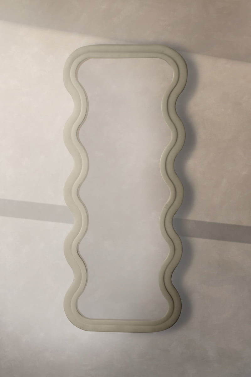 The Onde Mirror Large, featuring a tall rectangular shape and wavy velvet frame, hangs on a plain wall in soft natural light, adding contemporary sophistication to the space.