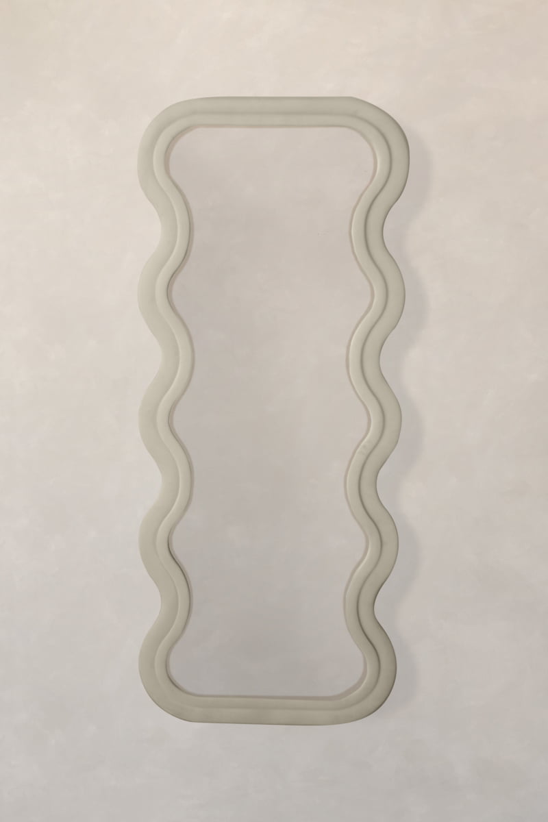 The Onde Mirror Large, featuring a tall rectangular shape and a wavy velvet frame, hangs on a light wall. Its soft, undulating design brings contemporary sophistication and playful modern style to your space.