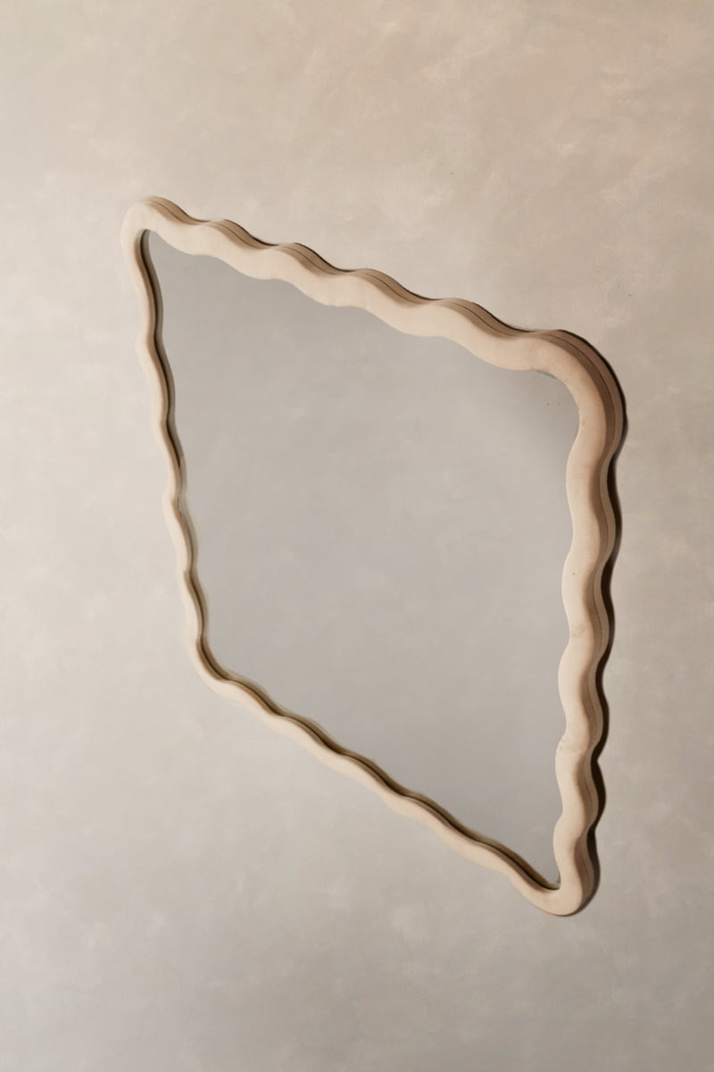 The Onde Mirror Small is a wall-mounted mirror with an irregular, wavy wooden frame and soft, organic lines—its modern minimalist design reflects a plain beige wall, making it ideal for contemporary interiors.
