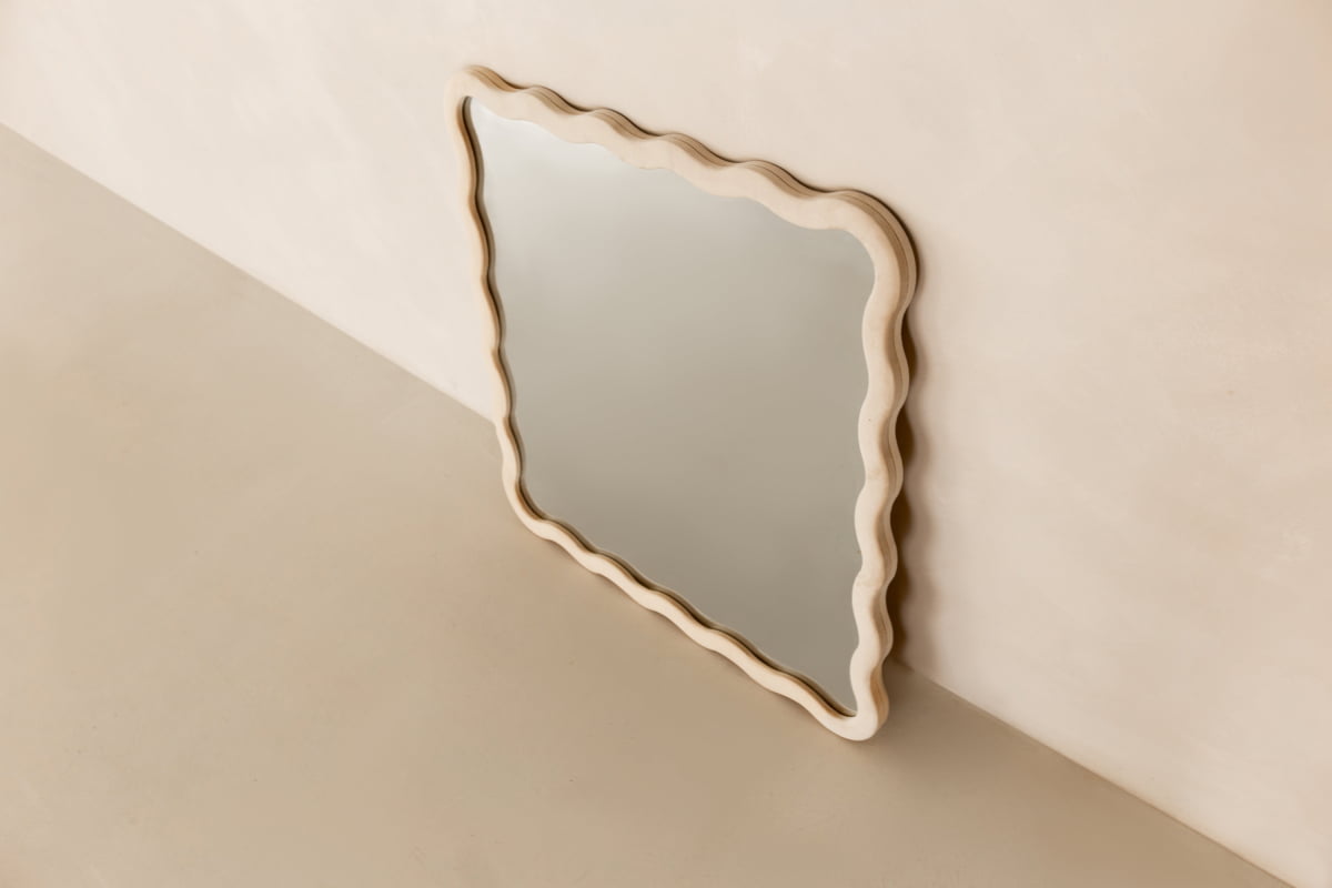 The Onde Mirror Small, featuring a rectangular shape and an irregular wavy wooden frame, leans against a beige wall on a light floor, adding a modern touch to the interior.