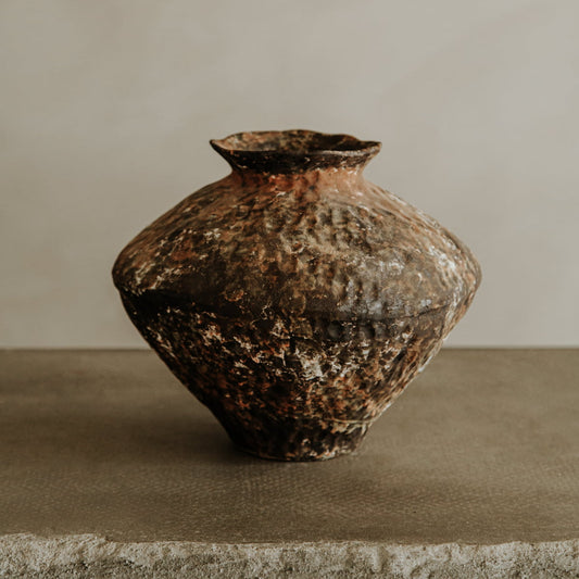The Oran ceramic vase, with its rustic texture and earthy brown tones, sits on a rough stone ledge against a neutral backdrop—perfect for adding warm character to your home decor.