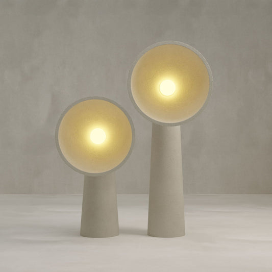 Two Orbis Short Floor Lamps—modern, cone-shaped designs with circular glowing yellow lights—stand on a neutral-toned surface, their sculptural artistry highlighted against a plain, textured beige background.
