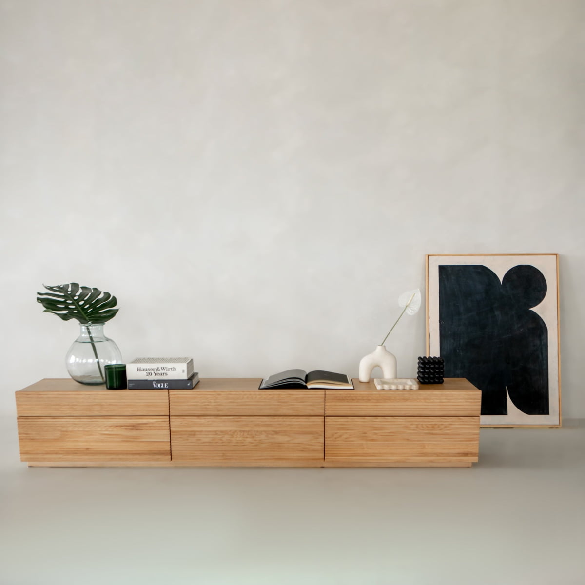 The Oster Media Console stands against a light gray wall, featuring discreet drawers and styled with a glass vase of green leaves, stacked books, an open book, a white abstract sculpture, a black candle, and framed abstract artwork.