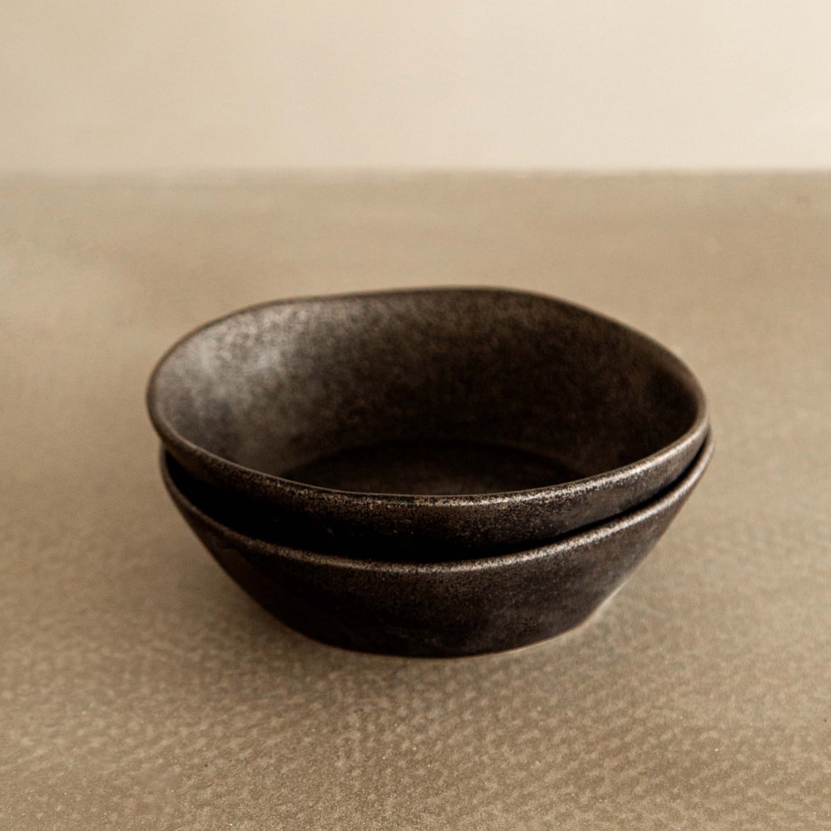 Two stacked Pebble Speckled Bowl Large ceramic bowls sit on a textured light brown surface with a plain off-white background.