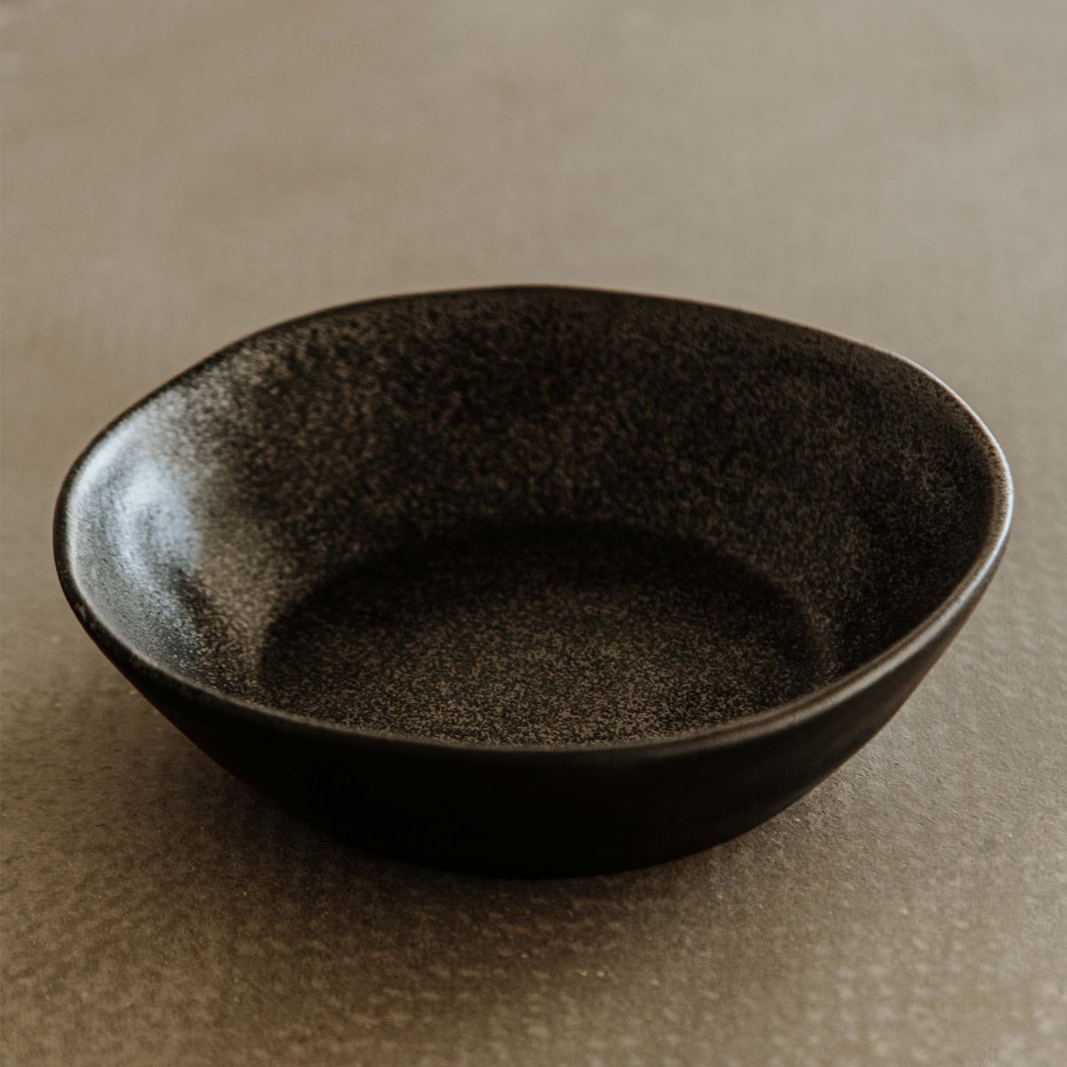 The Pebble Speckled Bowl Large is a dark, textured ceramic bowl with slightly uneven edges, shown empty on a light brown surface.