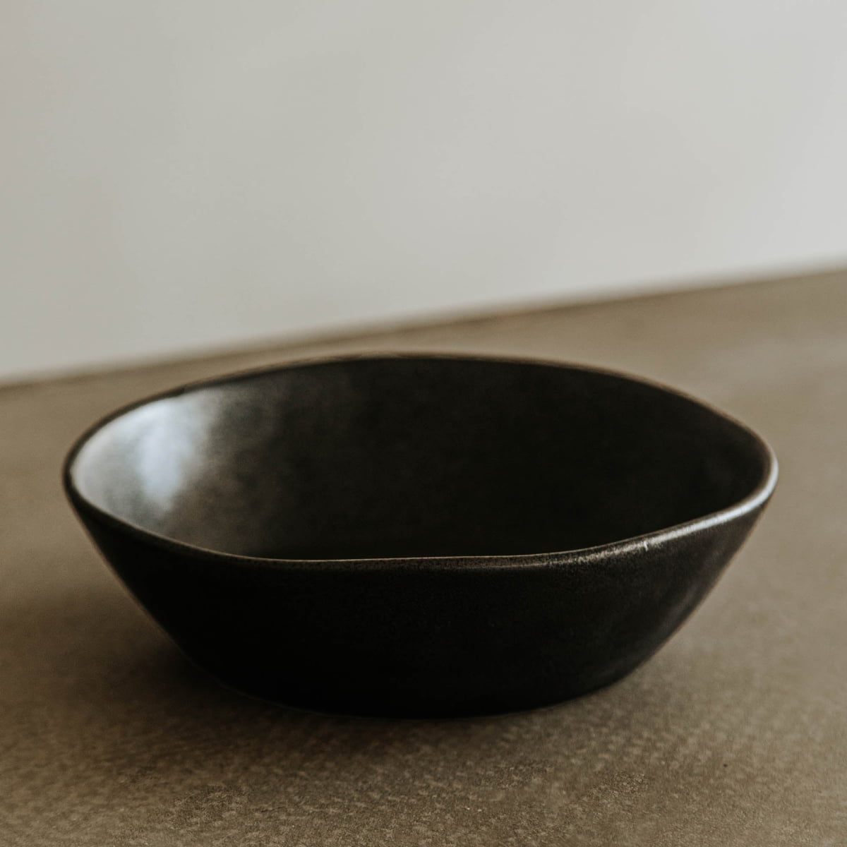 The Pebble Speckled Bowl Large, a black ceramic bowl with a matte finish, rests on a textured beige surface set against a plain, light background.