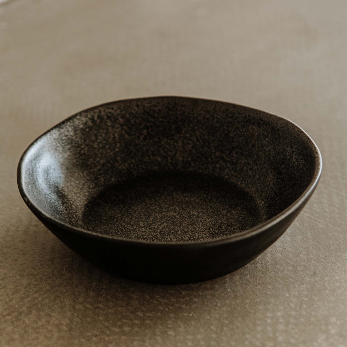 A Pebble Speckled Bowl Medium, black and round, sits empty on a textured light brown surface.