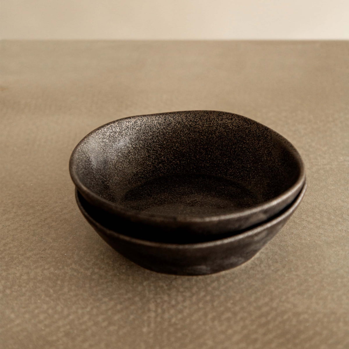 Two Pebble Speckled Bowl Mediums are stacked on a textured, light brown surface with a neutral background.