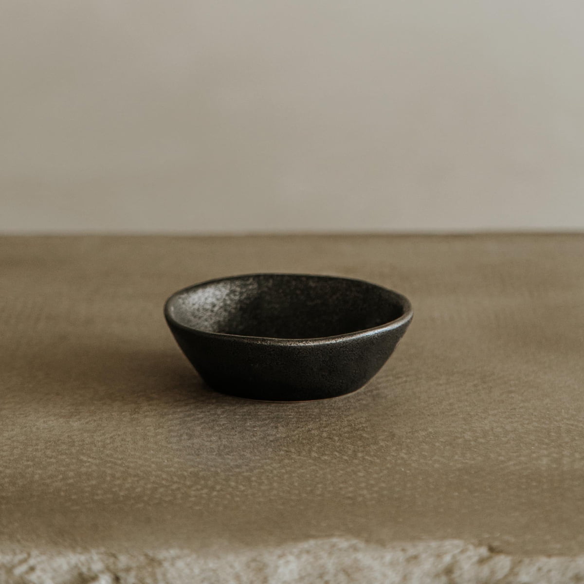 The Pebble Speckled Bowl Medium, a small black ceramic bowl, rests on a textured neutral surface with a soft beige background.