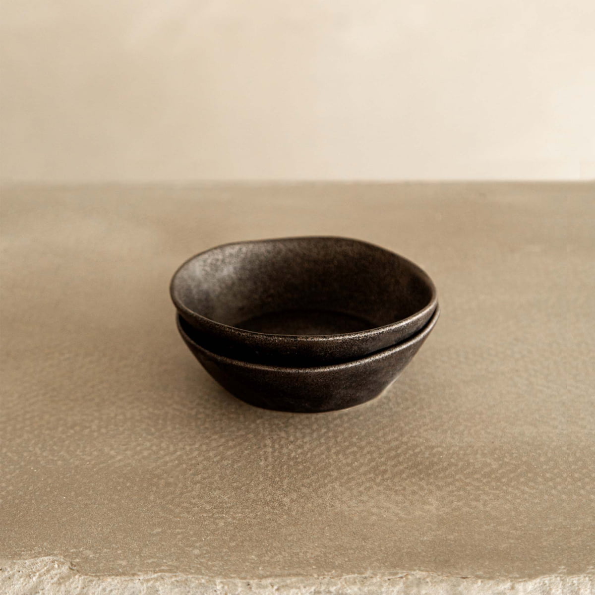 Two stacked Pebble Speckled Bowl Medium ceramic bowls, round and dark, sit on a textured beige surface with a neutral, blurred background.