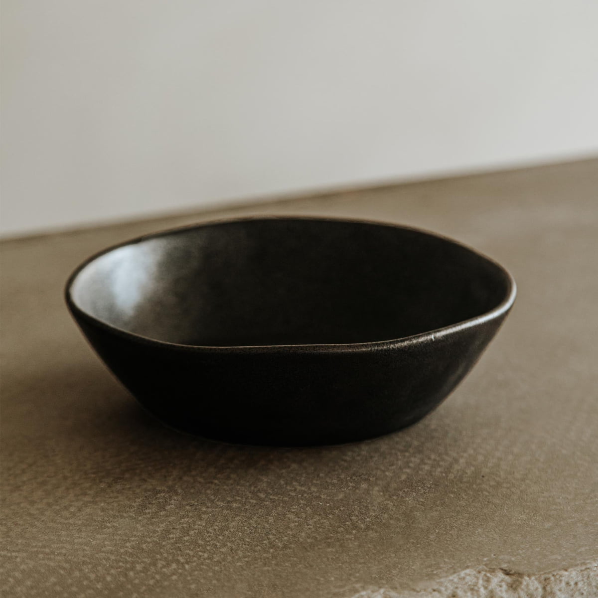 The Pebble Speckled Bowl Medium, featuring a simple round design in dark ceramic, rests on a textured light brown surface with a neutral background.