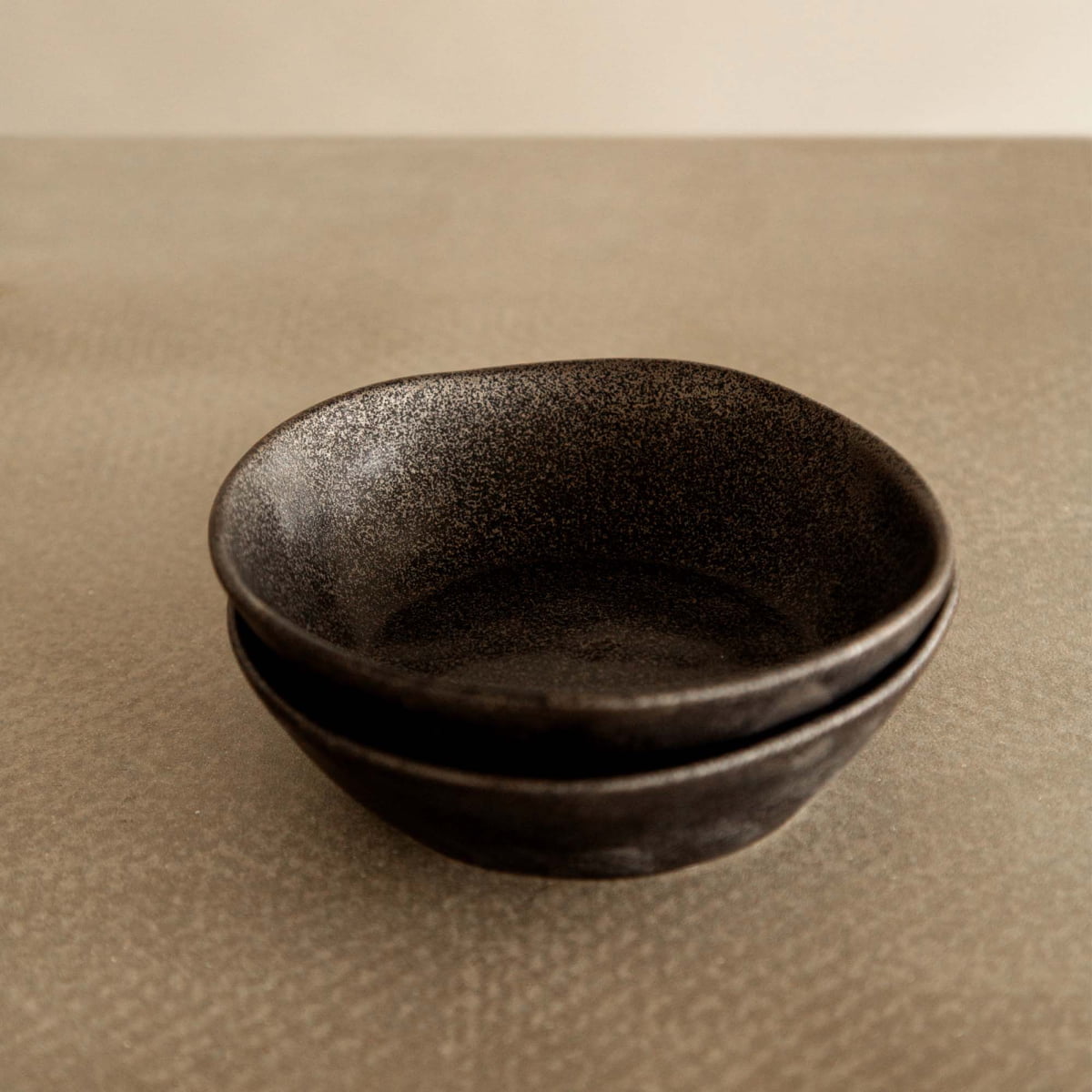 Two Pebble Speckled Bowl Small ceramic bowls are stacked together on a textured beige surface with a neutral background.