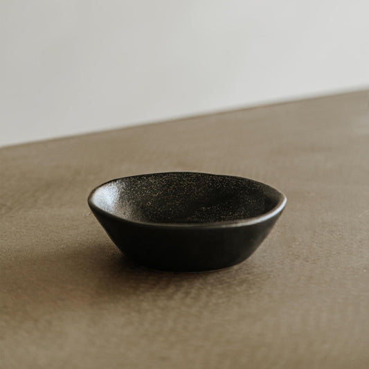 The Pebble Speckled Bowl Small, in black ceramic, rests on a textured brown surface against a neutral, blurred background.
