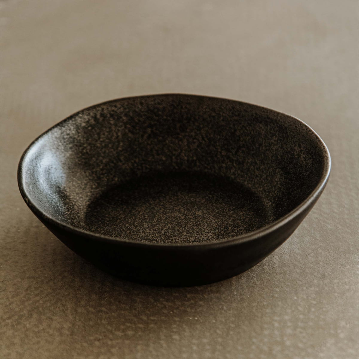 The Pebble Speckled Bowl Small, a black, round ceramic bowl with a textured finish, sits empty on a smooth, light brown surface.