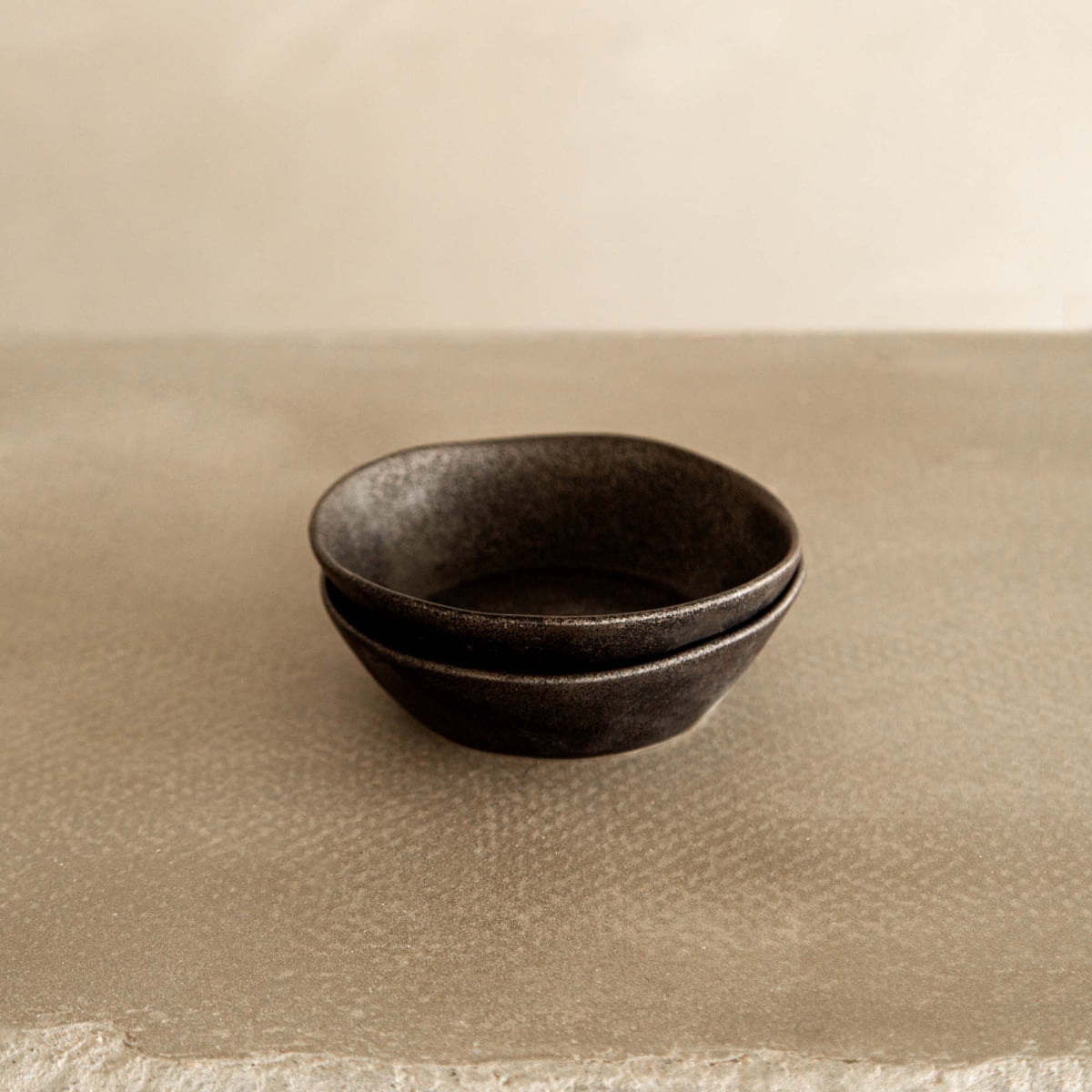 Two Pebble Speckled Bowl Small ceramic bowls are stacked on a textured beige surface, set against a soft, neutral background for a minimalistic and calming look.