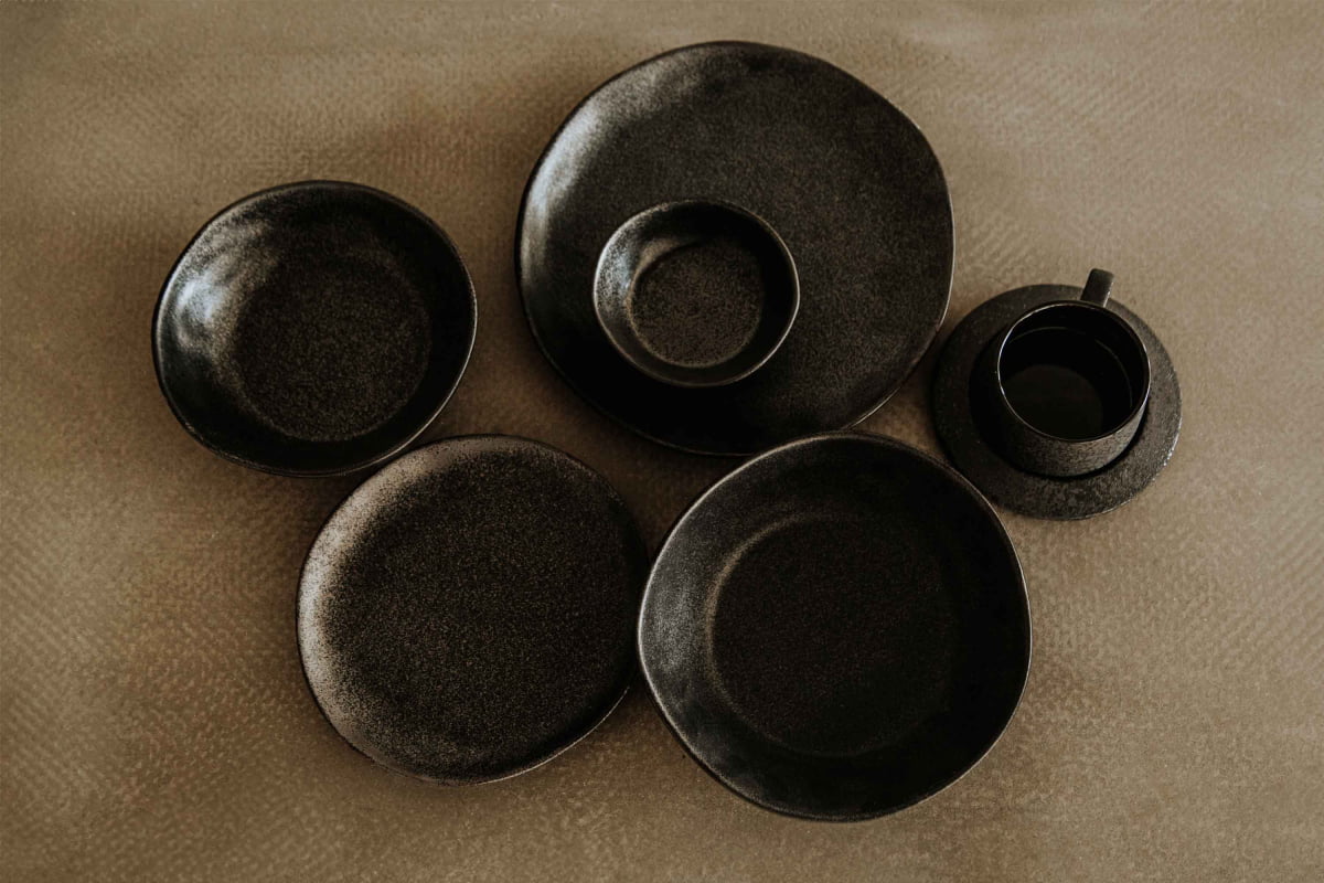 A Pebble Speckled Dish Small, along with four matte black ceramic dishes and a cup with saucer, are arranged on a textured beige surface. The varying sizes and shapes of the pieces create a visually balanced display.