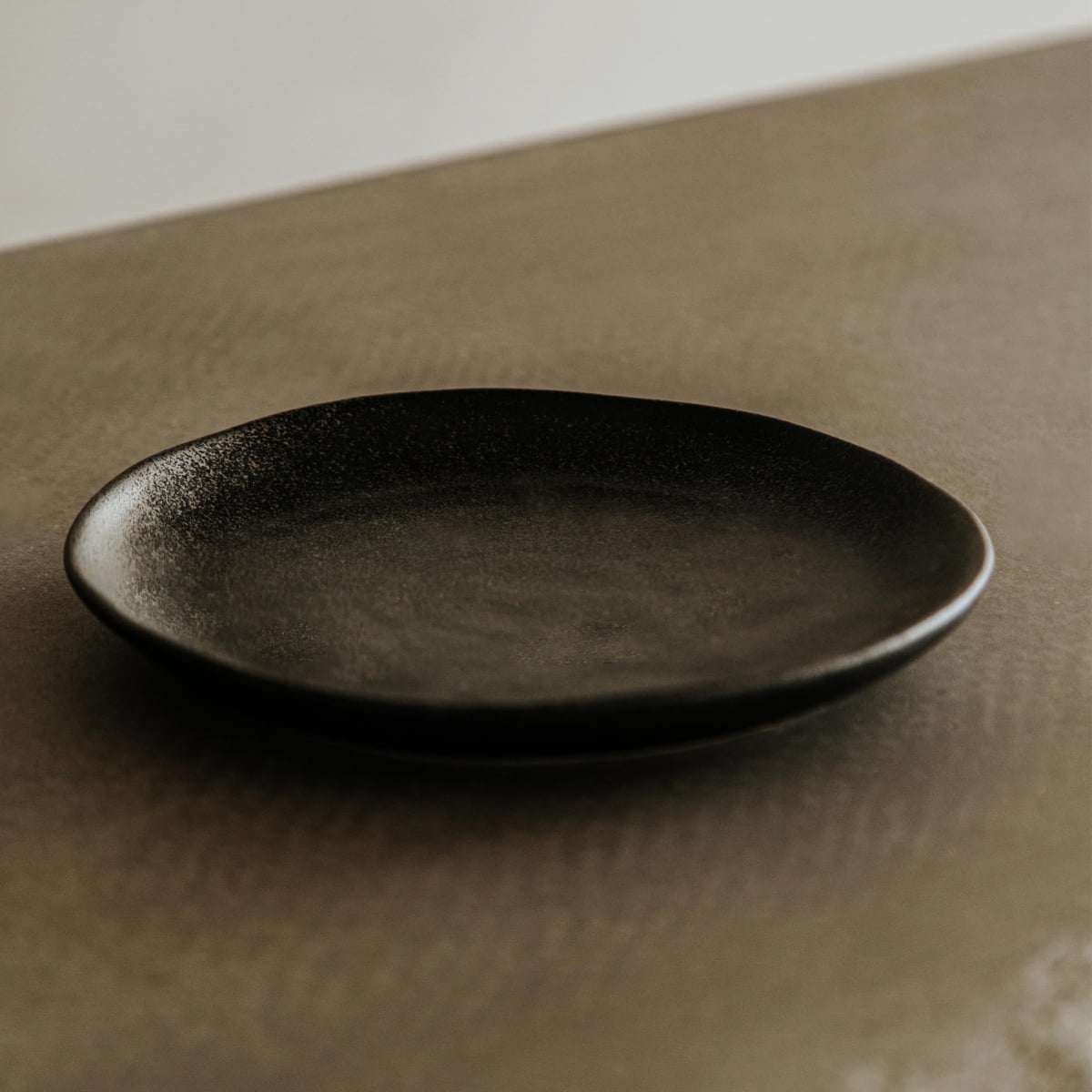 A single Pebble Speckled Dish Small, round and black, sits empty on a textured brown surface with a neutral background and soft lighting.