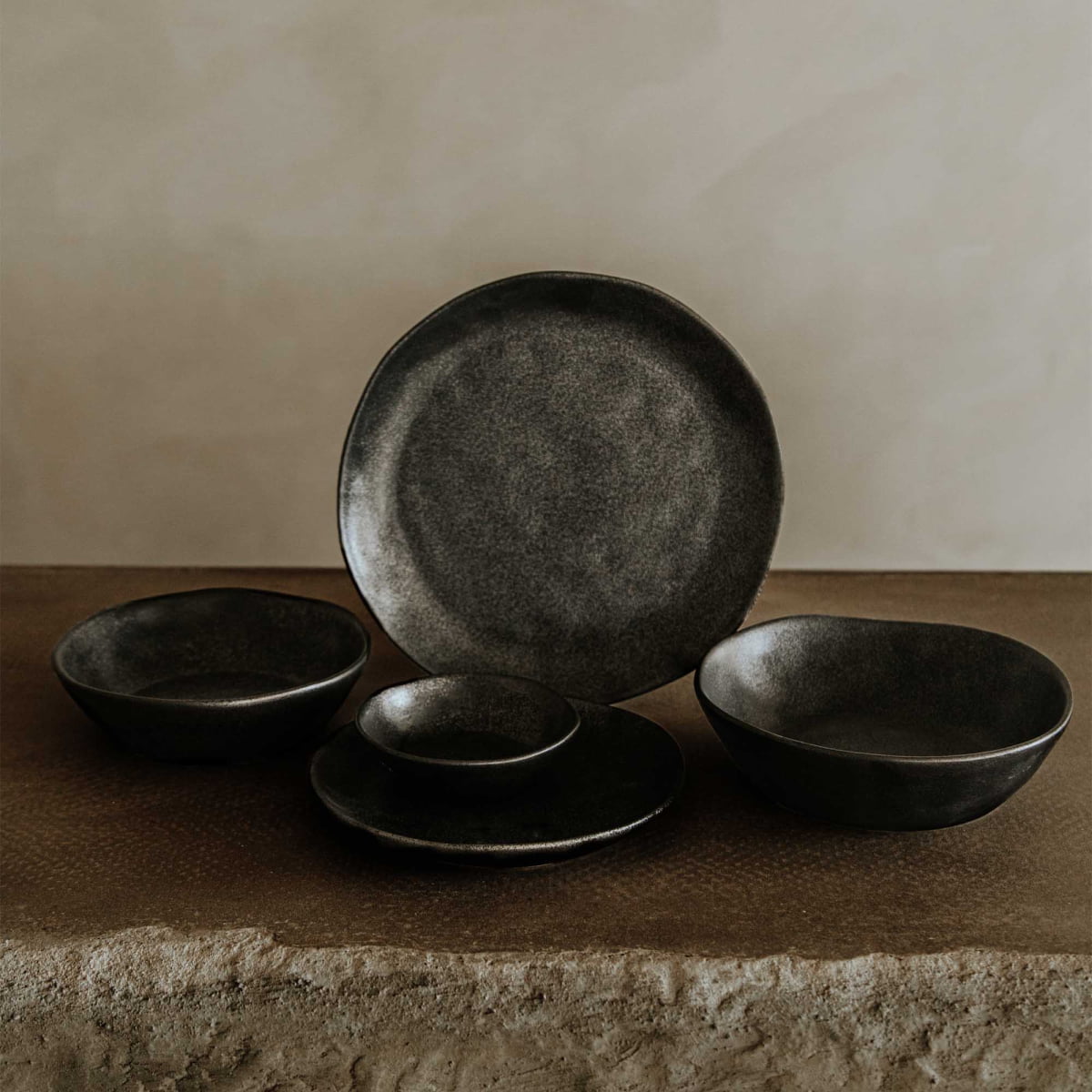 The Pebble Speckled Dish Small, a dark rustic ceramic piece, is displayed with similar bowls and plates on a textured stone surface against a neutral beige background.