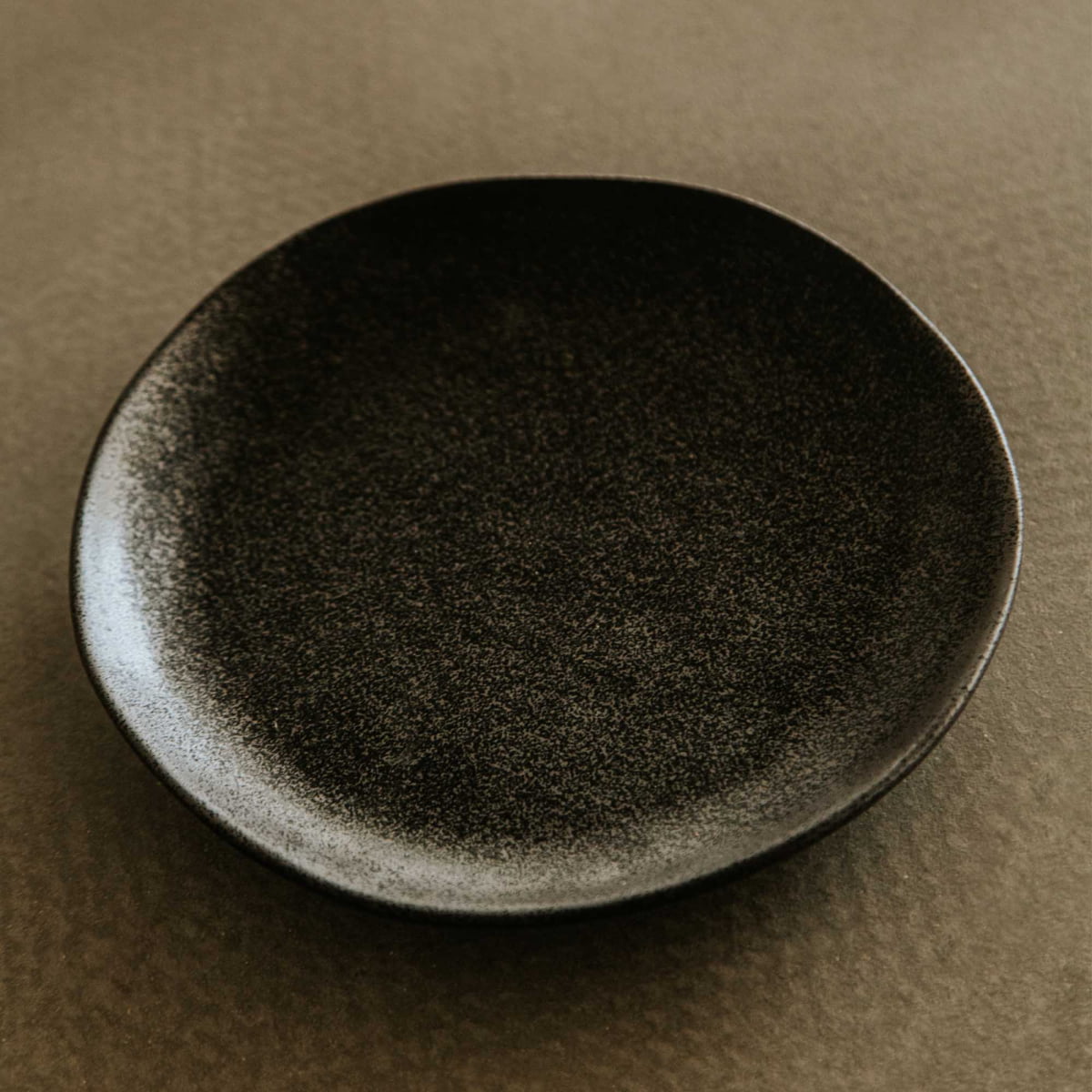 The Pebble Speckled Dish Small, a round ceramic plate with a slightly uneven rim and dark finish, rests empty on a textured brown surface.