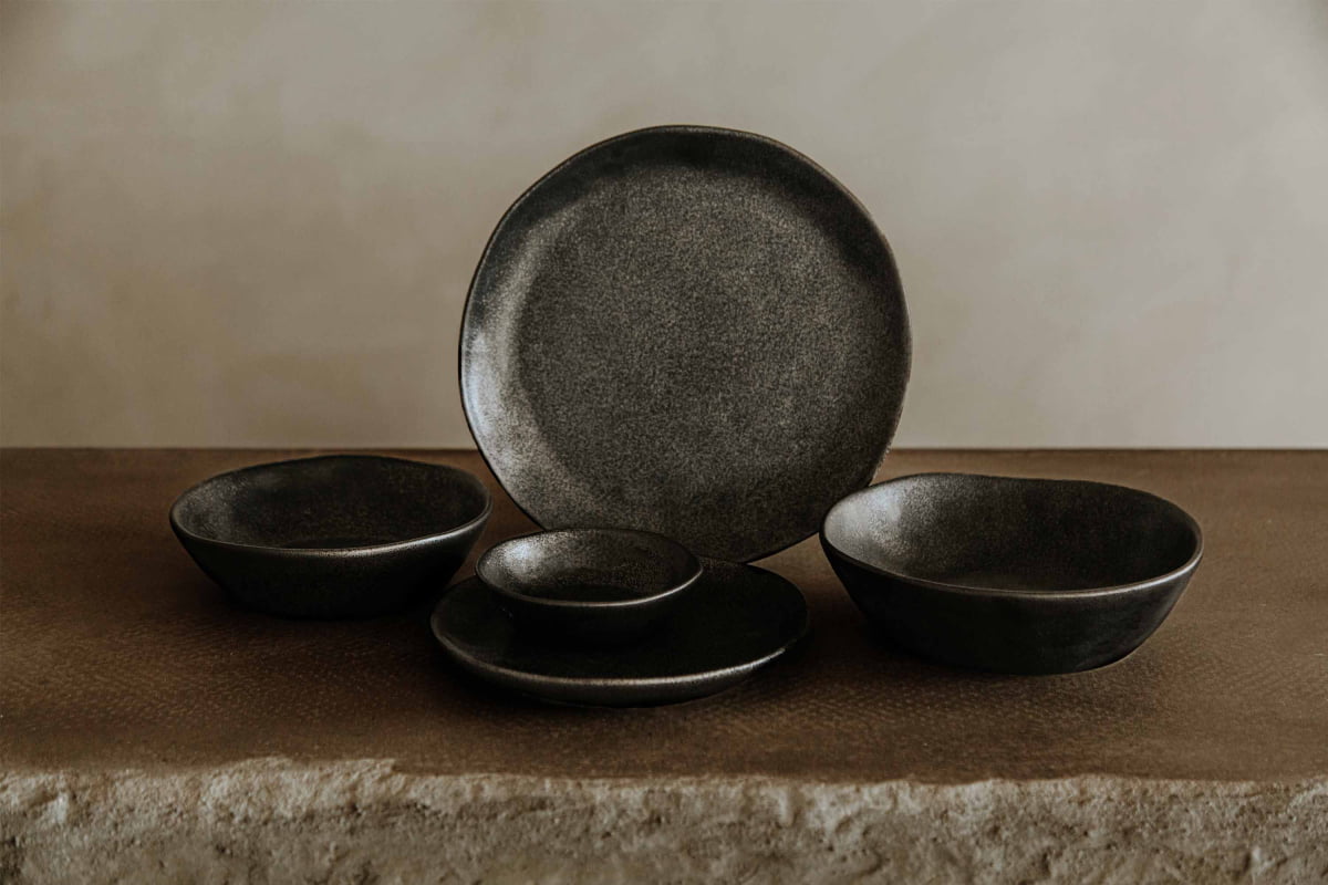 The Pebble Speckled Dish Small is a matte black ceramic dish, part of a set that also includes two bowls, one dinner plate, and one small plate, all arranged on a rustic brown surface against a neutral background.
