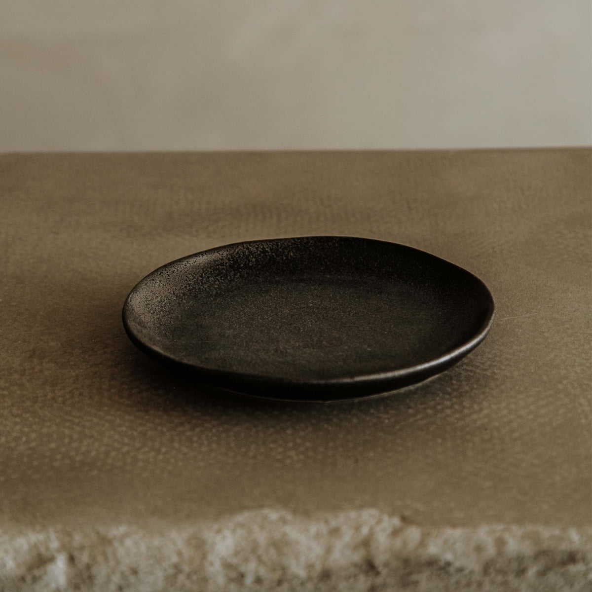 The Pebble Speckled Dish Small, a simple round black ceramic plate, rests on a textured beige stone surface against a neutral background.