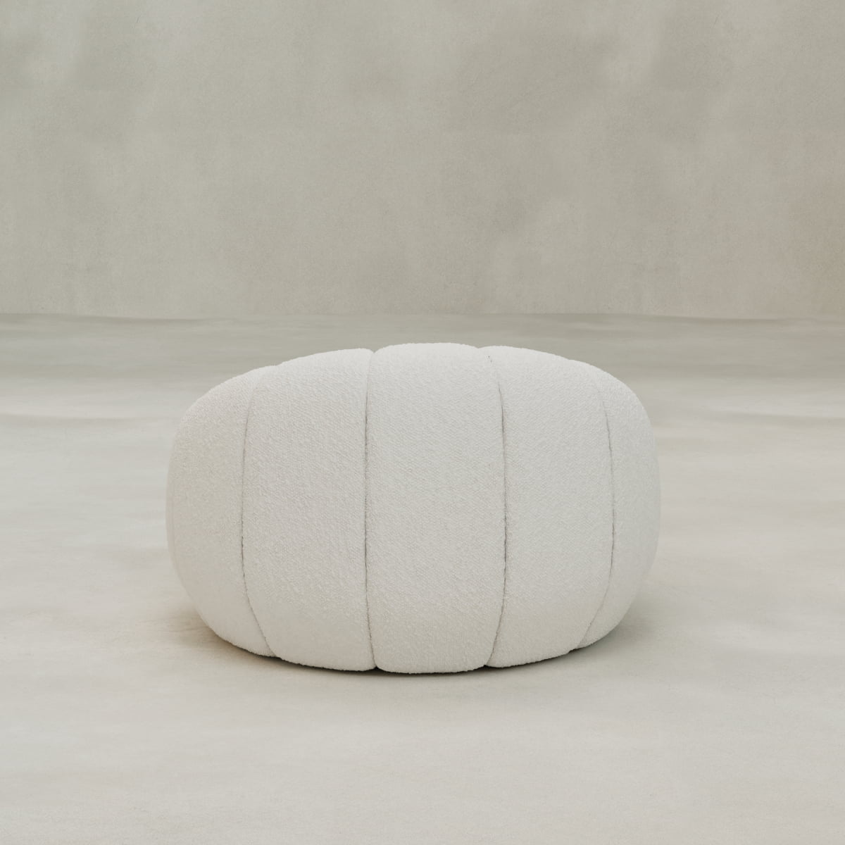 The Penelope Chair with Ottoman, featuring a round, white, tufted pouf in cozy bouclé with vertical seams, sits on a light floor against a neutral textured background.