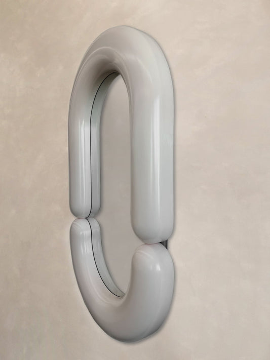 The Pitti Mirror Medium in White features a smooth, rounded, metallic chain link design mounted vertically on a light beige textured wall, bringing reflective elegance and artistic flair to any space.