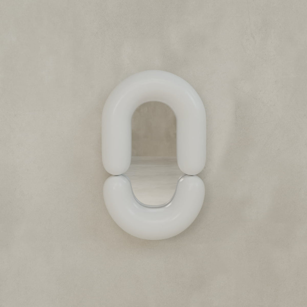 The Pitti Mirror In Medium features a minimalist, wall-mounted design with a reflective surface and a rounded white frame shaped like an incomplete oval, shown here against a beige textured wall.