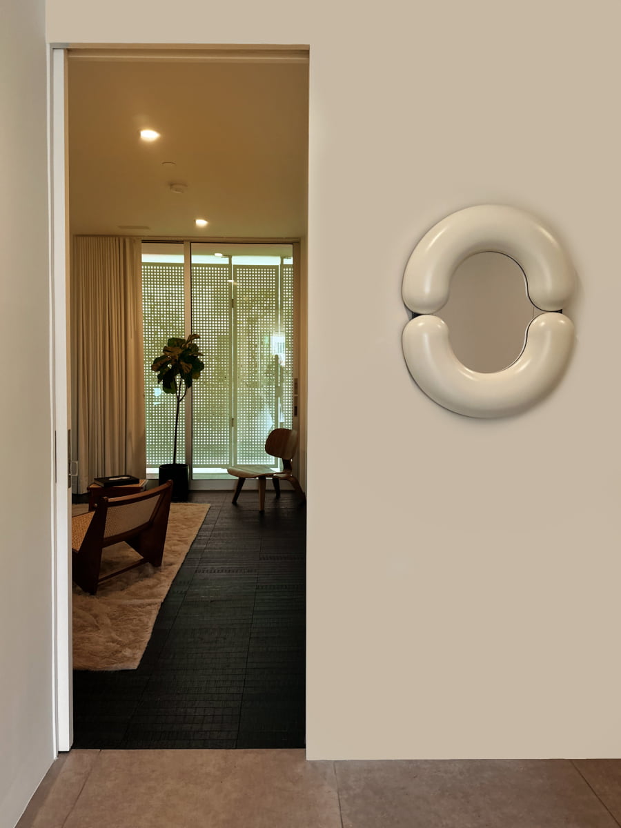 A modern, minimal living room is seen through an open door, with a wooden chair, low table, indoor plant, and floor-to-ceiling windows. The right wall features the Pitti Mirror Small In White, adding versatile décor with its reflective surface.