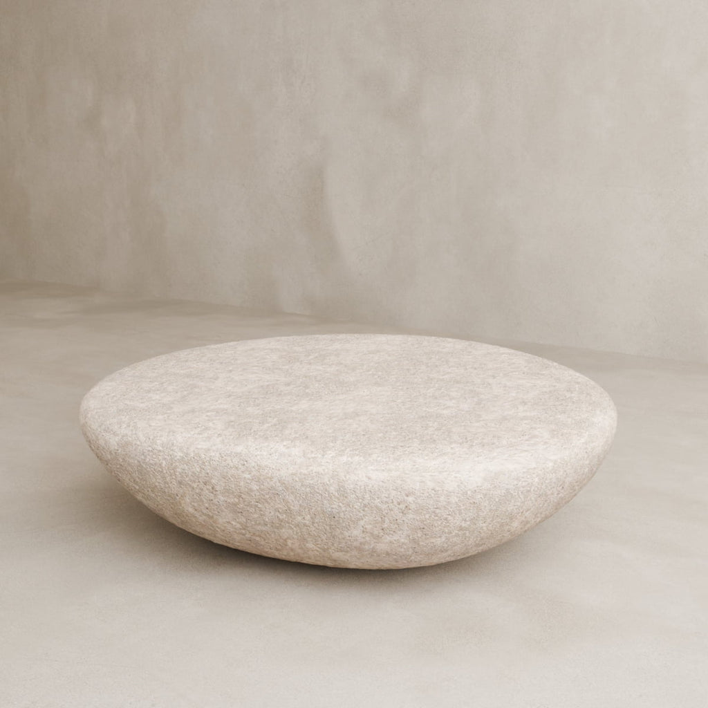 The Pluto Coffee Table, with its round white form and minimalist design, rests on a neutral floor by a textured wall, serving as a chic centerpiece that elevates the room’s modern aesthetic.